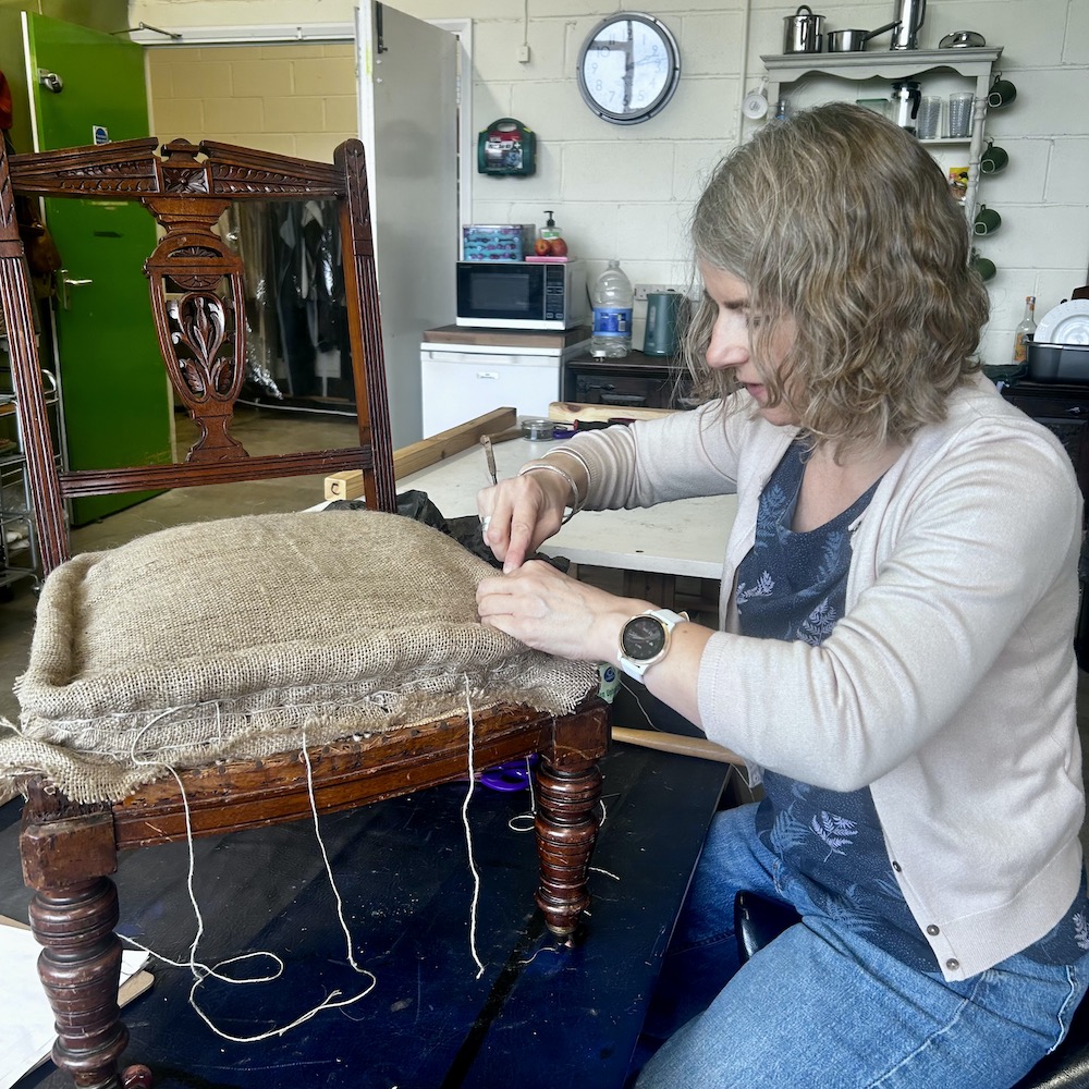 Student shaping and stitching traditional seat padding onto a wooden chair frame during a hands-on upholstery course.